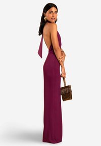 Vestido ajustado de cuello halter en burdeos oscuro con la espalda baja. Presenta un detalle atado y un bolso de mano dorado con lentejuelas y flecos.