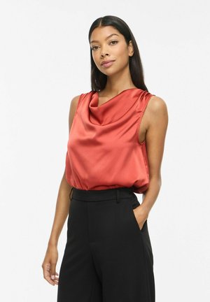Femme aux longs cheveux foncés portant un haut en satin rouge sans manches et un pantalon noir taille haute, debout avec une main dans la poche.