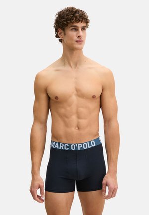 Männliches Modell mit lockigem Haar, trägt schwarze Marc O’Polo Boxershorts, steht vor einem einfarbigen hellgrauen Hintergrund.