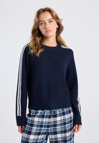 Navy sweater with ribbed cuffs and hem, featuring white stripes along the shoulders. Paired with blue and white plaid pants.