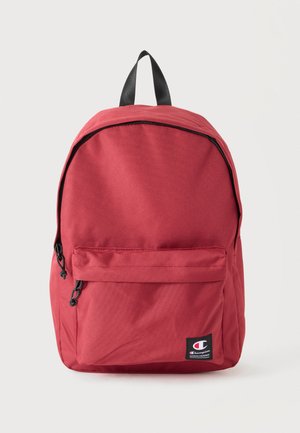 Sac à dos Champion rouge avec fermeture éclair noire, poche avant, boucle de poignée noire et étiquette avec le logo Champion sur un fond neutre.
