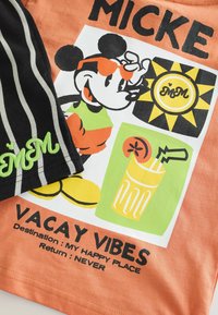 Orange bomulds t-shirt med en tegneseriefigur iført solbriller, en drik-grafik og tekst, der siger "VACAY VIBES", samt sorte stribede shorts.