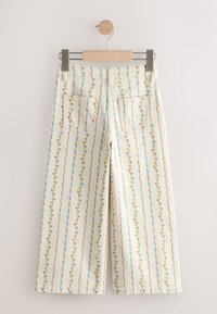 Pantalon crème à jambes larges avec des rayures florales multicolores verticales et deux poches plaquées au dos, suspendu sur un cintre en bois.