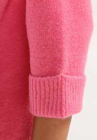 Manica di un maglione rosa a maglia con polsino a costine, texture morbida e design senza cuciture. Il materiale sembra caldo e accogliente, con un colore vivace.