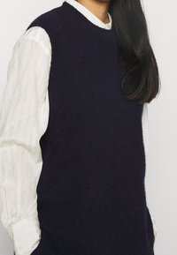 Stickad ärmlös tröja i mörk marinblå ull, med rund halsringning och ribbade kanter, bärs över en vit strukturerad skjorta med ett subtilt mönster.