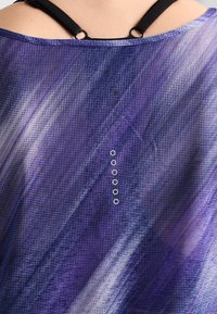 Close-up van een paars patroonstof met kleine metalen oogjes, gedragen over zwarte bandjes op de bovenrug van een persoon.