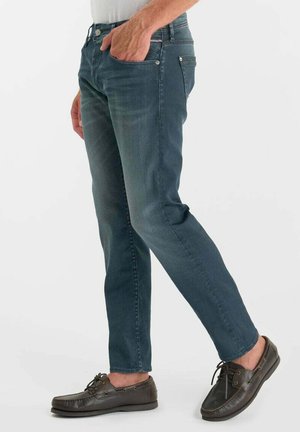Man draagt donkerblauwe slim-fit jeans en bruine leren veter loafers, staat met één hand in zijn zak tegen een effen achtergrond.