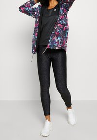 Kleurvolle bloemen-ritsjack, zwarte, aansluitende leggings met textuur, zwart T-shirt en witte sportieve schoenen. Het jack heeft zakken en trekkoorden.