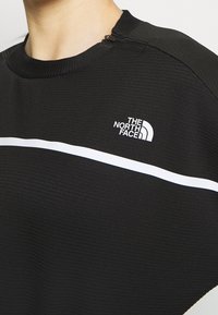 Primo piano di una persona che indossa un top North Face nero con texture, caratterizzato da una striscia orizzontale bianca e una zip vicino al colletto.