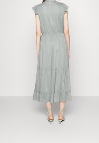 Kvinna klädd i en blekgrön metallisk midiklänning med volangärmar och flerlagad kjol, kombinerad med ljusgröna snörda klackskor med kittenheel.