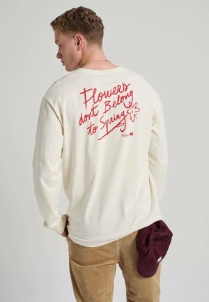 Mand iført cremefarvet langærmet skjorte med rød tekst "Flowers don't Belong to Spring" og holder en mørkerød kasket bag ryggen.