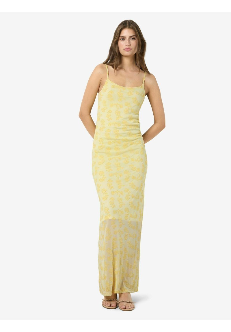 Femme debout les mains dans le dos, portant une longue robe nuisette jaune à fleurs et des sandales sur fond blanc.