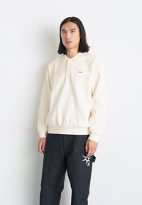 Sweatshirt polo de culoare crem, cu guler și manșete ribbrate. Prezintă un mic logo pe piept. Purtați-l cu pantaloni negri care au un accent de design.