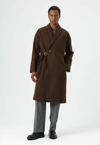 Bruine wollen jas met een reverskraag, sluiting met één knoop en een riem met een zilveren gesp. Draagt over grijze broek en zwarte schoenen.