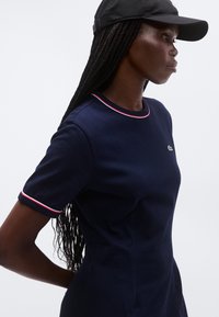 Γυναίκα με μακριά πλεξούδα, φορεί μπλε κοντομάνικη μπλούζα με το λογότυπο Croc και κόκκινη και λευκή διακόσμηση, καθώς και μαύρο καπέλο, κοιτάζει προς τα δεξιά σε λευκό φόντο.