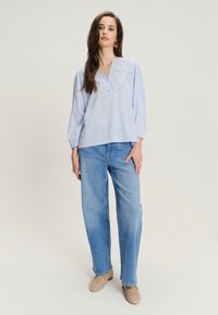 Blusa azzurro chiaro a maniche lunghe con scollatura a V, caratterizzata da un delicato ricamo floreale sul petto. Indossata con jeans blu larghi e scarpe beige.