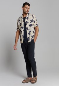 Korte mouwen bloeshemd in crème met blauwe en gele prints, gecombineerd met een donkere broek en bruine sandaaltjes met gespen.