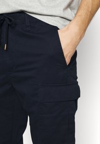 Close-up of a person wearing navy blue cargo pants with a side pocket and a hand placed inside the front pocket.