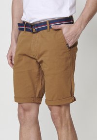 Man i bruna uppvikta shorts och ett blått tygbälte med röd och vit rand, hand i fickan, mot en neutral bakgrund.