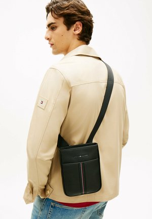 Jonge man met een beige jas en blauwe spijkerbroek, met een zwarte schoudertas met een verticale rood-wit-blauwe streep.