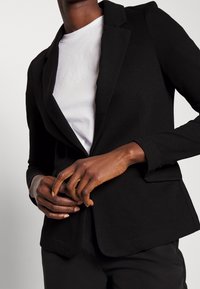 Blazer noir avec une coupe ajustée, des revers structurés et des poches avant, porté avec un t-shirt blanc en dessous.