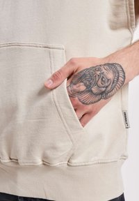 Beige Kapuzenpullover aus weicher Baumwolle, mit einer Fronttasche und geripptem Saum. Zeigt ein detailliertes Tattoo auf einer teilweise sichtbaren Hand.