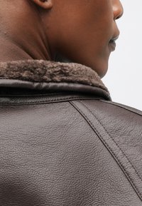 Καφέ δερμάτινο μπουφάν με μαλακό γιακά από γούνα, υφή επιφάνειας και ορατές ραφές, εστιάζοντας στην πλάτη του ρούχου.