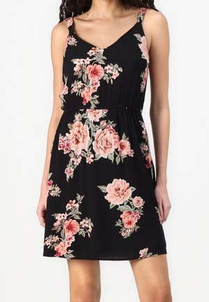 Femme portant une robe sans manches noire avec des motifs floraux roses et corail, ajustée à la taille, sur un fond clair uni.