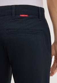 Granatowe spodnie w wzór jodełki z tylną kieszenią. Czerwona metka "JOOP! JEANS" jest przyszyta nad kieszenią, co podkreśla fakturowany materiał.