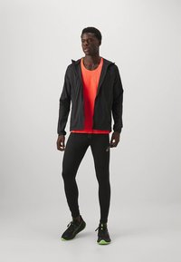 Veste de sport noire, débardeur orange néon, leggings noirs, et chaussures noires avec des accents verts. Tissu lisse avec un design respirant.