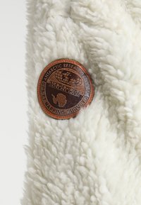 Vitt fluffigt tyg med en texturerad yta, med en cirkulär brun läderlapp med texten "ANTARKTISK FORSKNINGSPROGRAM" och en karta över Antarktis.