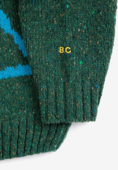 Manche de pull tricoté vert foncé tacheté avec des lettres jaunes brodées "BC" et une partie d'un motif bleu sur le tissu.