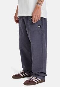 Donkergrijze cargobroek met een ontspannen pasvorm, voorzien van zijzakken en een klein zwart logoplaatje. Gecombineerd met bruine en witte sneakers.