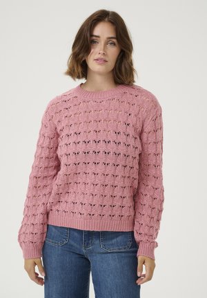 Pull en tricot rose avec un motif en maille ouverte texturée et des poignets côtelés, associé à un jean bleu, sur fond blanc uni.