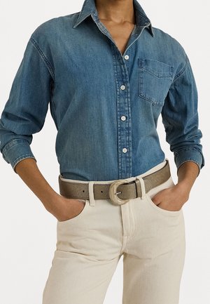 Persona con camisa de mezclilla azul abotonada metida en pantalones beige con un cinturón marrón claro, manos en los bolsillos, cuello abierto.