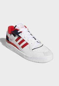 Baskets blanches avec des accents rouges et noirs, arborant trois bandes rouges Adidas et le logo sur la languette et le talon, montrées sous un léger angle.
