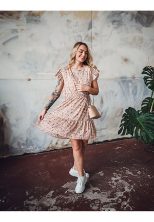 Blumendruckkleid mit kurzen Ärmeln, gerafftem Bund und gestuftem Rock in Hellrosa, kombiniert mit weißen Sneakers und einer kleinen Handtasche.