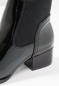 Bottine noire avec une finition brillante en vernis, panneaux élastiques sur les côtés, petit talon carré et coutures soignées.