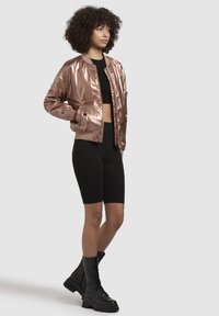 Metalická růžová bomber bunda, zkrácený design, žebrované manžety, nošena s černým crop topem a černými cyklistickými šortkami, doplněná chunky černými šněrovacími botami.