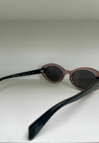 Lunettes de soleil ovales avec une monture translucide rose et des verres foncés. Les branches sont entièrement noires, affichant un logo et des détails sur le modèle.