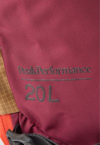 Tecido bordô com um acabamento texturizado, apresentando um logótipo da PeakPerformance e "20L" em cinza. Acentuado por uma área padronizada em castanho claro e acessórios em preto.