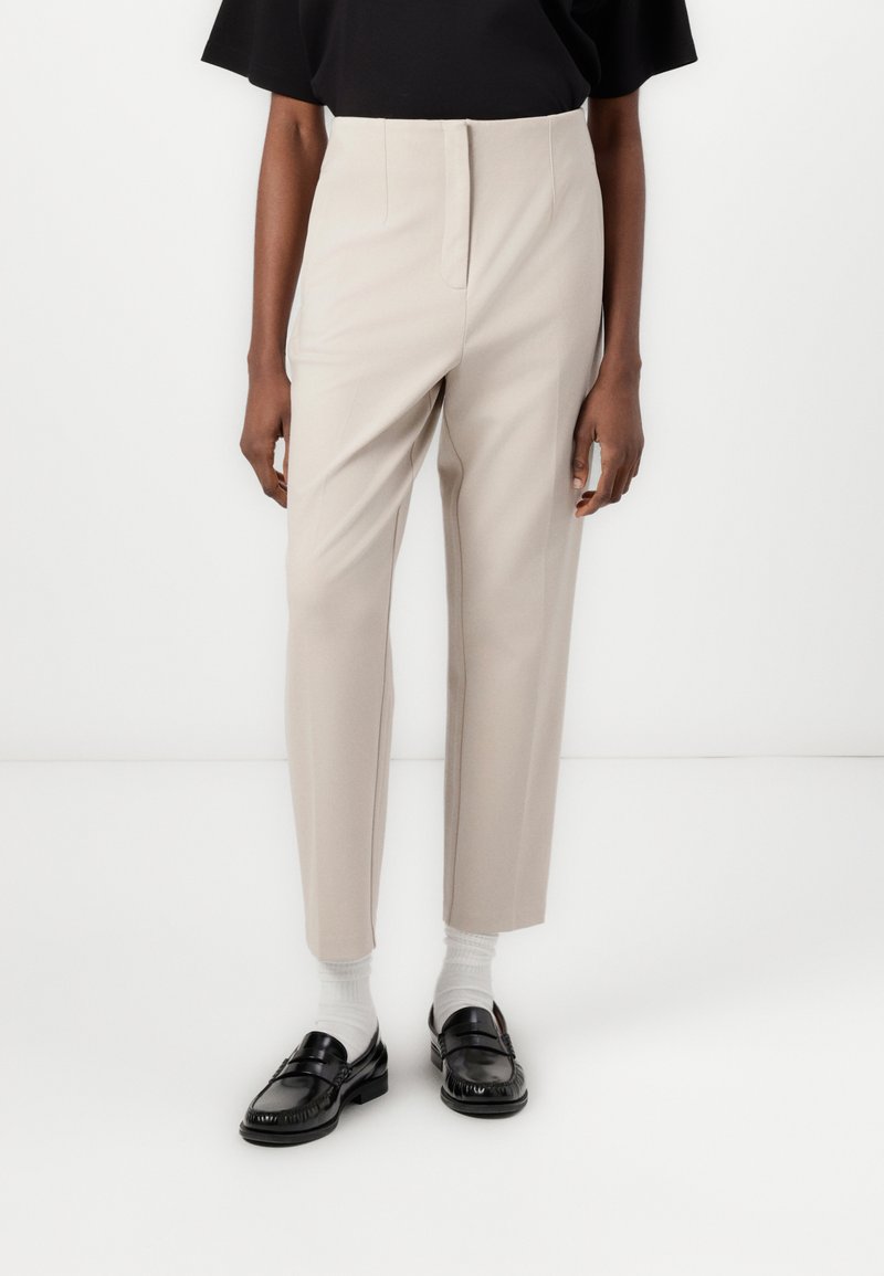 Light beige tailored trousers with a high waist, straight legs, and front pleats. Paired with black loafers and white socks.