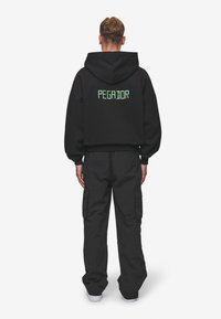 Black hooded sweatshirt with "PEGADOR" in neon green lettering on the back, paired with loose-fitting black cargo pants.