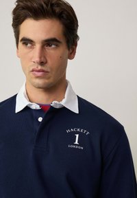 Mornarska polo majica s bijelim ovratnikom, s natpisom "HACKETT" i brojem "1" otisnutim u bijeloj boji. Mekana tekstura, kopčanje s gumbima, standardni kroj.