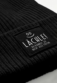 Black knitted beanie featuring a textured fabric and a rectangular black patch with white text and a smiley face illustration.