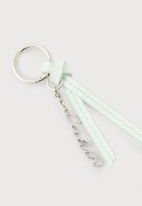 Ασημένιο μπρελόκ Calvin Klein με δύο λουριά από ανοικτό πράσινο δέρμα και ένα κρεμαστό κόσμημα Calvin σε γραφή χειρόγραφου, προσαρτημένο με αλυσίδα.