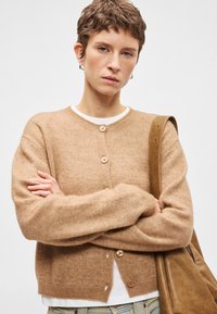 Ανοιχτό καφέ μαλλί κάρντιγκ με στρογγυλή λαιμόκοψη και τέσσερα κουμπιά, με μακριά μανίκια και χαλαρή εφαρμογή. Ορατό ζευγάρι ανοιχτό μπλε τζιν.