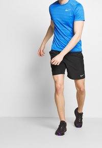 Blå, kortärmad träningströja med horisontella ränder, kombinerad med svarta shorts och svarta sneakers med lila detaljer.