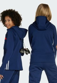 Tamno plava sportska dukserica s kapuljačom i crvenim detaljem, koja ima teksturirani prednji dizajn, u kombinaciji s odgovarajućim hlačama i drži kopačke za nogomet.