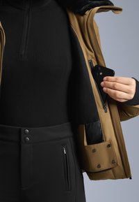 Brown jacket with a black zippered inner pocket. A black fitted top and high-waisted black pants are also visible. Smooth texture and sleek design.